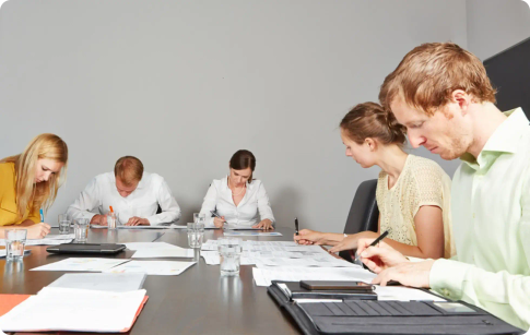 Several people sit around a table, all of them are looking down and writing on paper.