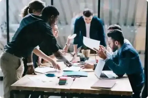 A team works together around a table covered with documents and laptops.