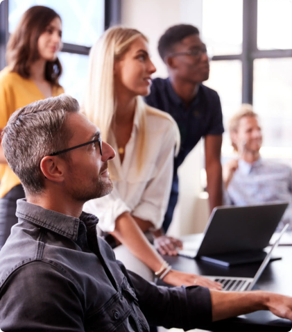 A group of people gathered around a desk with their laptops listens intently to someone else in the room.