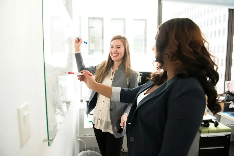 Two women at work collaboratively using a whiteboard, smiling and laughing together.